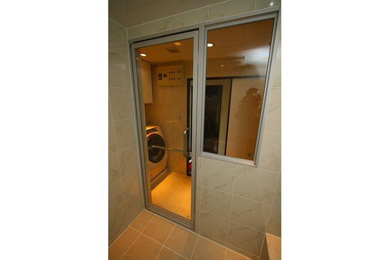 浴室から洗面室を映した写真です。ガラスのステンレス製の取っ手がオシャレです。床のタイルは、LIXIL社製のサーモタイルです。サーモタイルは、寒い冬も、あまり冷っとしないんです。